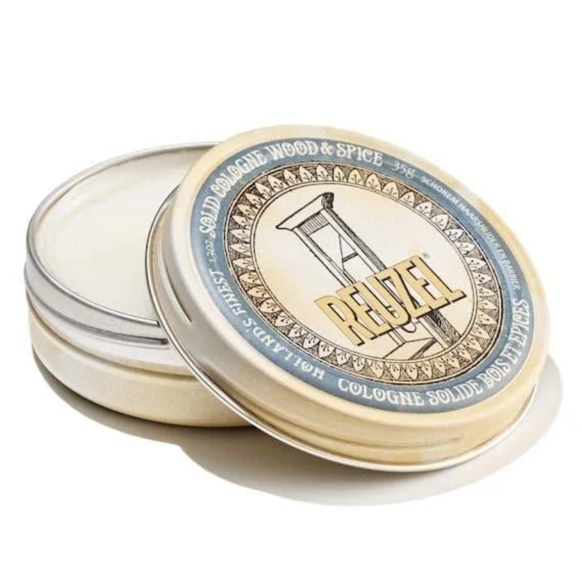 A round metal tin of Reuzel Cologne Solid 100% in the Wood & Spice scent, with the lid propped open to reveal the cream-colored solid inside. The label features a vintage-style illustration of a lighthouse and the brand name 'REGZEL' in gold.