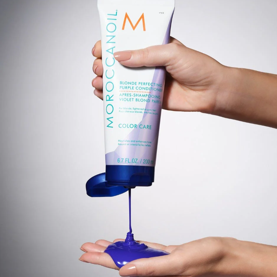 Hand holding a tube of Moroccanoil product with blue substance being dispensed onto another hand.