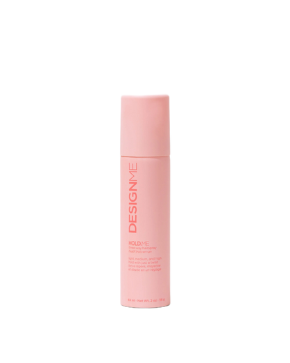 A tall, cylindrical pink bottle of Design Me Hold+ve hair hold spray, shown against a solid white background.