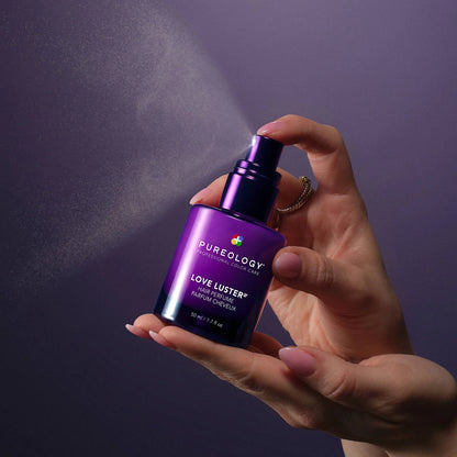 Hand holding a purple Pureology hair product with a spray action against a dark background