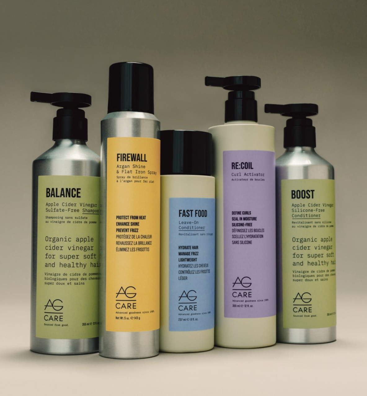Five AG Care haircare bottles in a row against a neutral beige background. The bottles are silver and black with labels in yellow, orange, dark blue, cream, and silver.