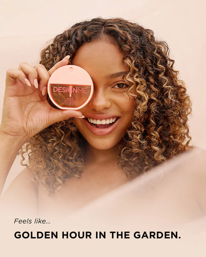 A smiling woman with curly hair holds a round makeup compact over one eye. The compact lid shows the brand name DESIGN ME.