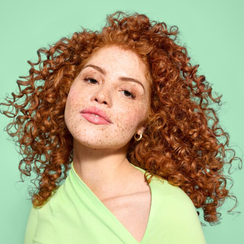 A close-up portrait of a woman with voluminous, tightly curled auburn hair and freckles, wearing a light green top against a solid mint green background.