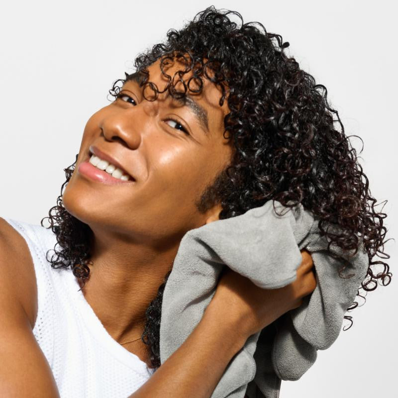 A smiling person with curly dark hair drying their face and neck with a gray towel, wearing a white sleeveless top against a light gray background.