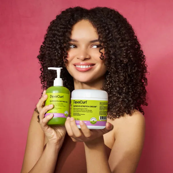 A smiling woman with voluminous dark curly hair holds two DyeaCurl products against a solid pink background: a green Quinoa Creaming Gel pump bottle and a white Quinoa Stretch Cream jar.
