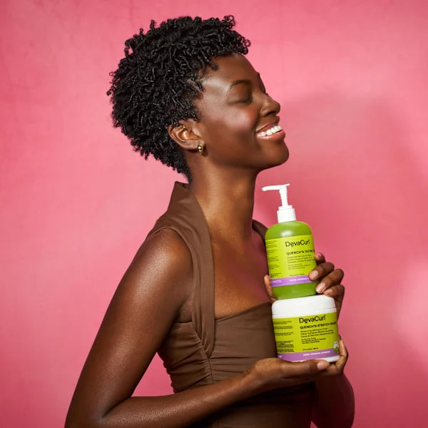 A smiling woman with short curly black hair holds two DevaCurl hair care bottles against a solid pink background. She is wearing a brown sleeveless top, and the bottles are lime green and white with purple accents.