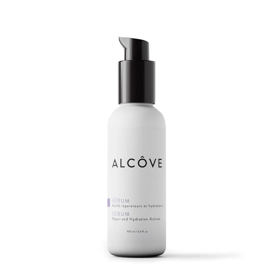 A bottle of Alcôve Serum on a clean white background. The bottle is a frosted light gray with a black pump dispenser and features the brand name 'ALCÔVE' in black text.