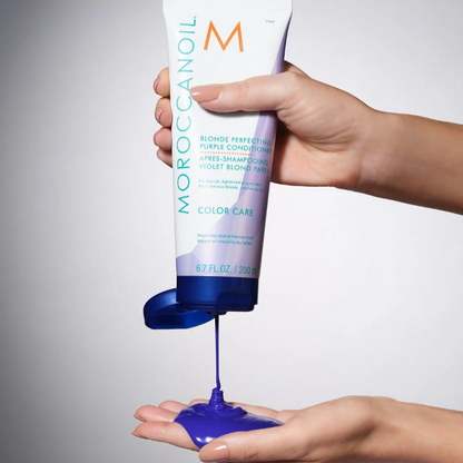 Hand holding a tube of Moroccanoil product with blue substance being dispensed onto another hand.