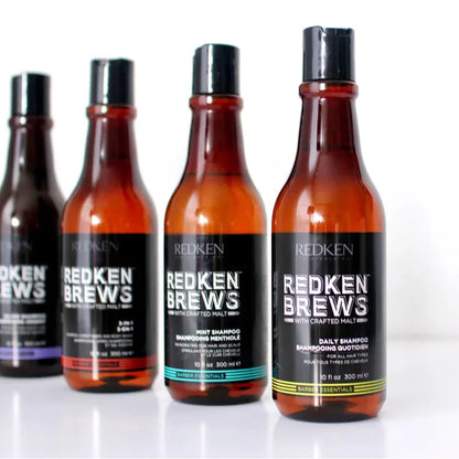 Four Redken haircare bottles in amber glass with black caps and black labels, arranged in a row on a white reflective surface. The front bottle is labeled Redken Brews Daily Shampoo, and the others include Mint Shampoo, Infused Hair Wash, and another dark bottle.