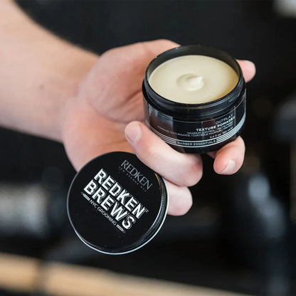 A hand holds an open black tin of Redken Color Styling Texture Reviver hair wax, showing the pale cream-colored product inside, with the black lid placed in front. The background is a dark, blurred environment.